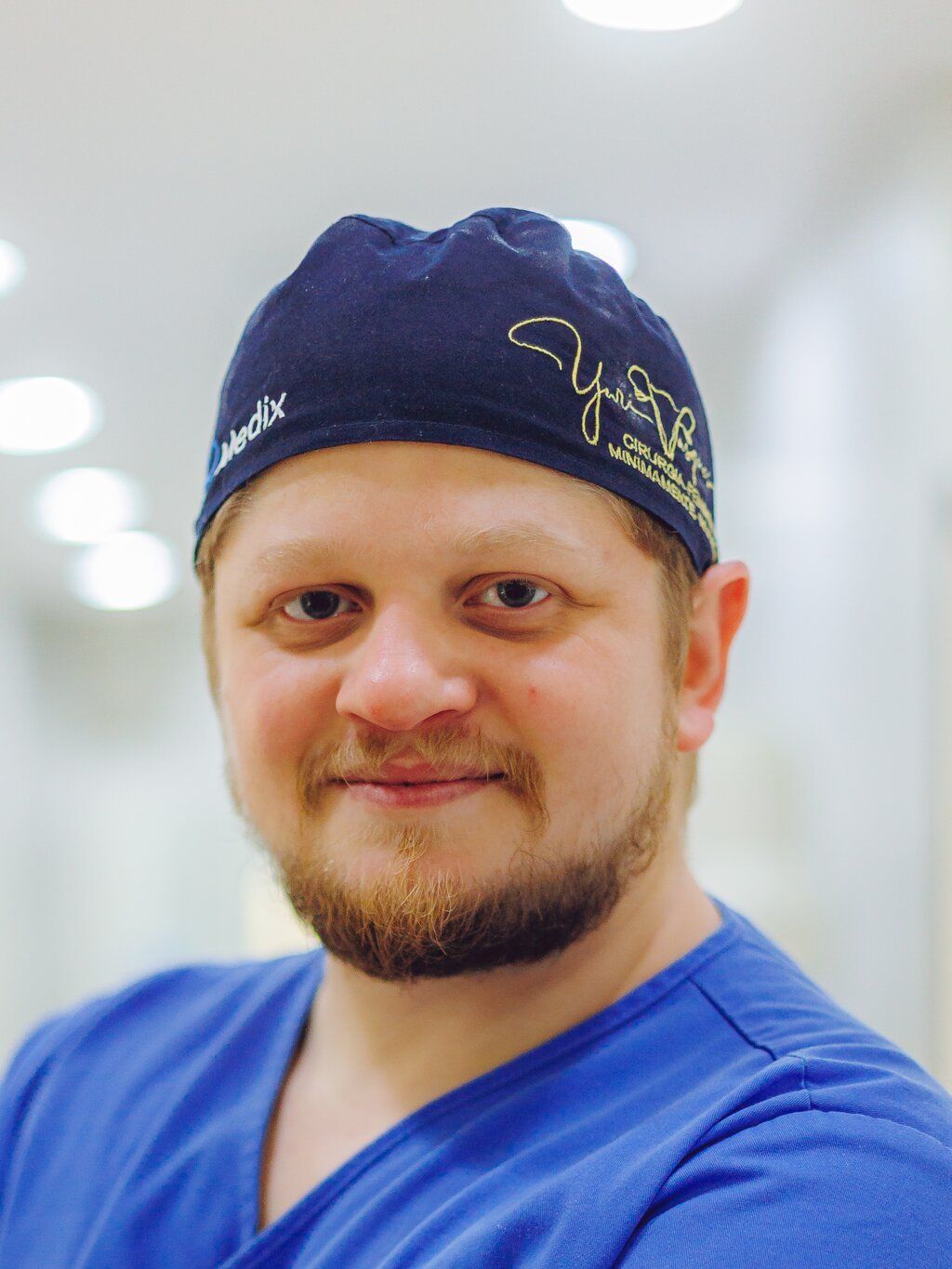 Dr. Yuri Vasques sorrindo, de pijama cirúrgico e touca, no corredor do centro cirúrgico