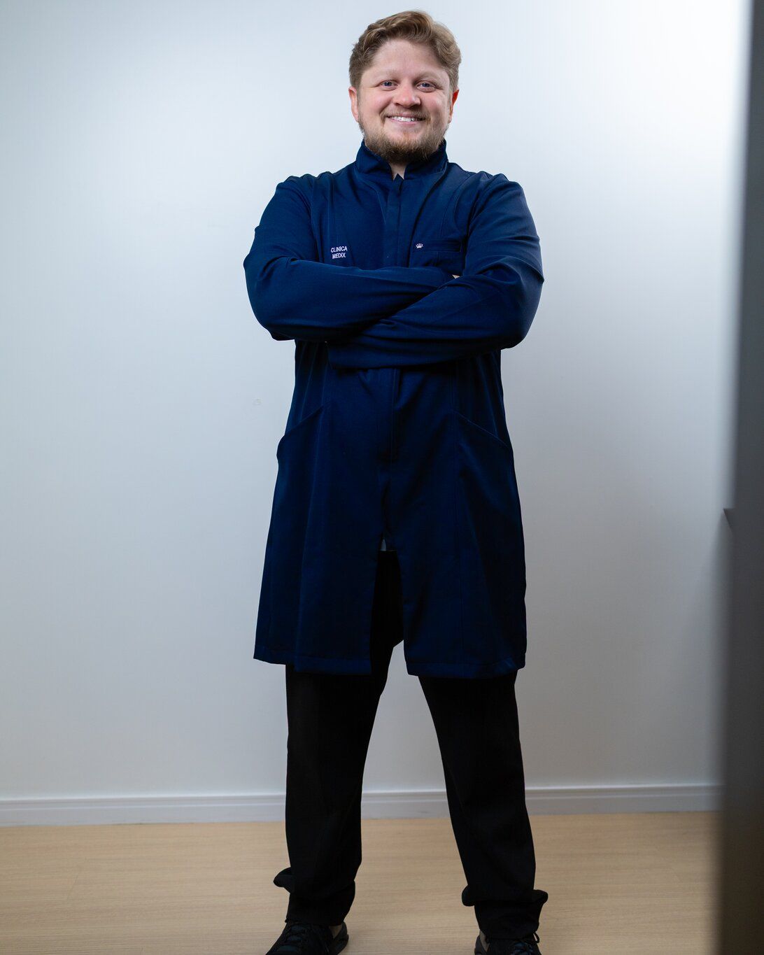 Dr. Yuri Vasques de jaleco azul e braços cruzados, de corpo inteiro