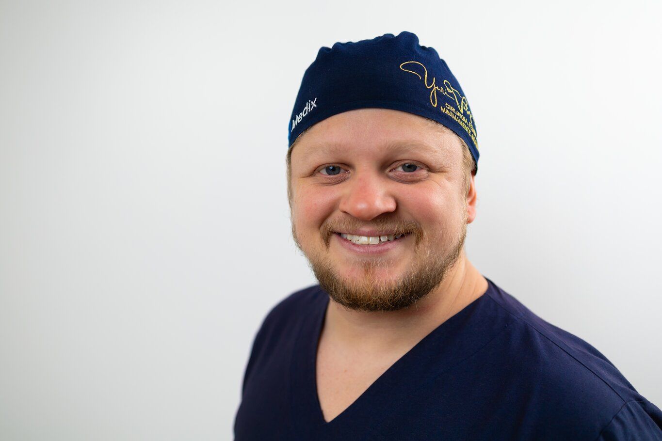 Dr. Yuri Vasques sorrindo de orelha a orelha, de pijama cirúrgico e touca com seu nome